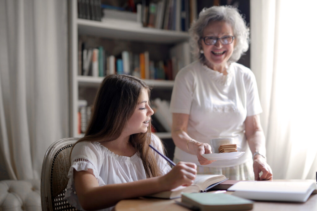 3768164-3768164 Cheerful grandmother in glasses and casual clothes smiling at camera while giving pastry on plate and mug of tasty beverage to joyful teenager sitting at table with books and exercise book and study in light cozy living room